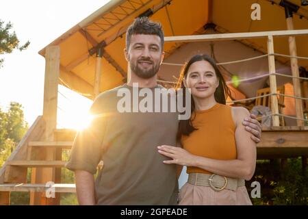 Glücklicher junger Mann, der seine Frau gegen das Glamping-Zelt umarmt, während er vor der Kamera steht Stockfoto