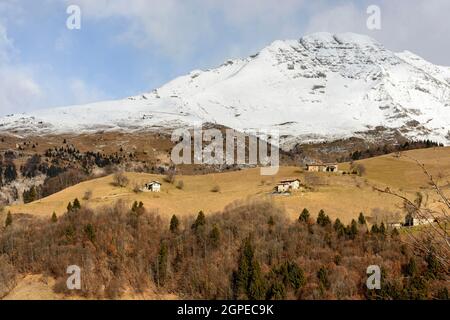 Landschaft mit Berghütten auf grünen Lichtungen unter dem schneebedeckten felsigen Hang des Arera Peak in Bergamo Berge in einem Winter mit wenig Schnee, in b geschossen Stockfoto