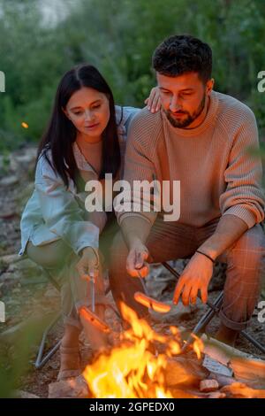 Junges liebevolles Paar, das Würstchen über dem Feuer braten und sich in natürlicher Umgebung entspannen kann Stockfoto