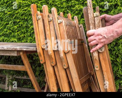 Nahaufnahme der Hand eines Mannes, der vier gefaltete Holzgarten-/Gartenstühle an einen alten Tisch stapelte, draußen bei herbstlicher Sonne. Stockfoto