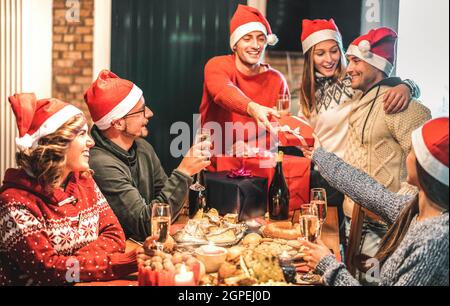 Freunde Gruppe trägt weihnachtsmütze geben einander Weihnachtsgeschenke - Champagner Wein Toast zu Hause Weihnachten Abendessen - Feiertagskonzept mit jungen Menschen Stockfoto