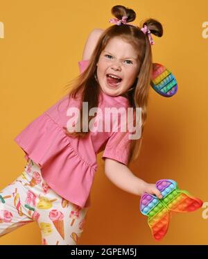 Frolic, aktive Mädchen in bunten Kleidern spielt mit zwei sensorischen Regenbogenfarben Spielzeug - Pop it in die Hände Stockfoto