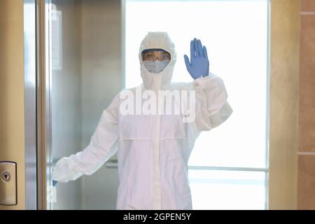 Frau in Schutzanzug und medizinische Maske mit Handschuhen steht im Aufzug Stockfoto