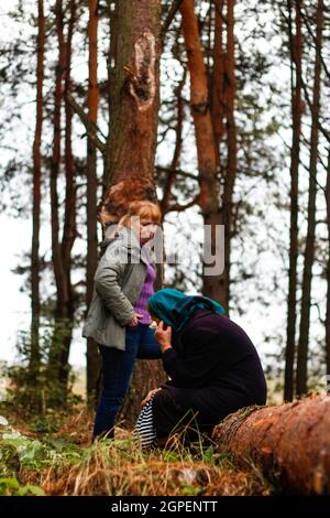 Unschärfe-Seitenansicht von zwei Frauen, die im Pinienwald spazieren und auf einem Baumstamm sitzen. Freizeit- und Menschenkonzept, Mutter und Tochter im Herbstwald. Menschen Stockfoto