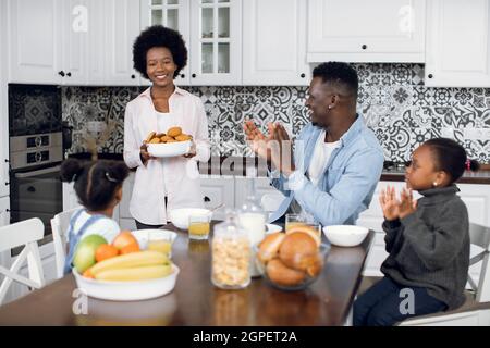 Schöne afrikanische Frau hält Teller mit handgefertigten Cookies, während sie auf der modernen Küche steht. Mann und zwei Töchter sitzen zusammen am Tisch und Stockfoto