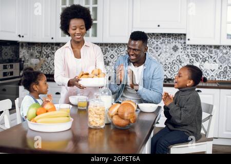 Liebenswert afroamerikanischen Frauen serviert auf dem Tisch leckere hausgemachte Kekse für ihre schöne Familie. Zwei glückliche Töchter und ein gutaussehender Mann sitzen zusammen in der modernen Küche und klatschen die Hände Stockfoto