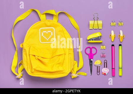 Zurück zur Schule, Bildungskonzept. Gelber Rucksack mit Schulbedarf - Notizbuch, Stifte, Clips, Schere isoliert auf violettem Hintergrund Draufsicht Copy Stockfoto