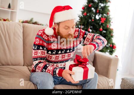 Foto Porträt bärtigen Mann Auspacken Weihnachten Geschenk-Box in Kopfbedeckung neugierig auf der Couch sitzen Stockfoto