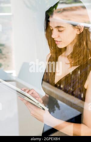 Die junge Frau in formellem Kleid arbeitet zu Hause auf ihrem Tablet, wie sie durch das Fenster gesehen wird Stockfoto