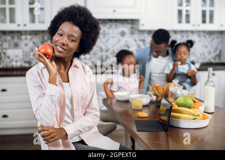 Angenehme afroamerikanische Frau, die lächelt und die Kamera anschaut, während sie in der Küche mit Apfel in den Händen sitzt. Ehemann und zwei Töchter spielen im Hintergrund. Häusliches Leben der glücklichen Familie. Stockfoto