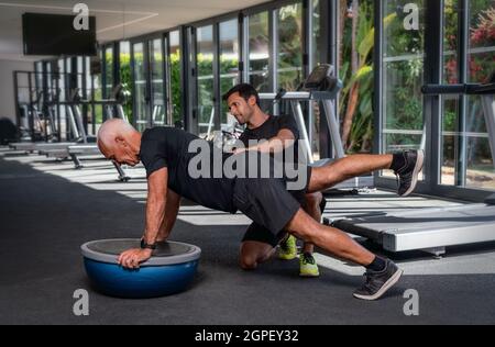 Ein älterer kaukasischer Mann, im Fitnessstudio, mit einem persönlichen Fitnesstrainer, in der Rehabilitation, macht Übungen für das Gleichgewicht auf einem Gymnastikball. Stockfoto