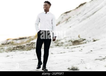 Schöner eleganter schwarzer Mann, in einem weißen Hemd, geht mit seinem Laptop, im Freien vor dem Hintergrund der weißen Kreide Felsen und Berge, Konzept fo Stockfoto