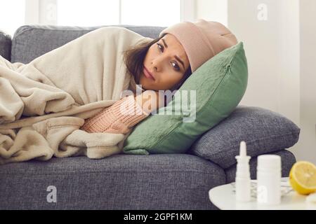 Junge unglückliche Frau in warmen Kleidern und Hut, die im Bett unter der Decke lag und sich krank fühlte Stockfoto