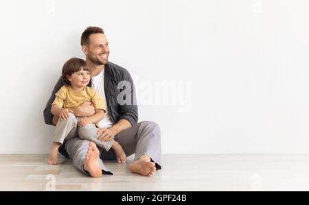 Glücklicher kaukasischer Vater, der einen kleinen Sohn trägt, zusammen auf dem Boden sitzt und auf einen leeren, weißen Hintergrund blickt Stockfoto
