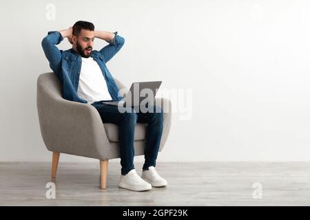 Schockierter junger arabischer Mann, der seinen Kopf in der Nähe eines Laptops schnappte und Fehler bei einem Online-Job-Projekt in der Nähe einer weißen Studiowand machte Stockfoto