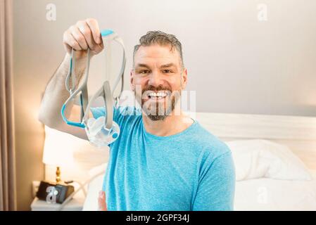 Reifer glücklicher Mann mit chronischen Atemproblemen erwägt die Verwendung CPAP-Maschine auf dem Bett im Schlafzimmer sitzen. Gesundheitswesen, CPAP, Schnarchen Konzept Stockfoto