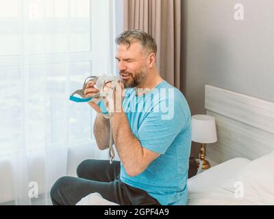 Reifer glücklicher Mann mit chronischen Atemproblemen erwägt die Verwendung CPAP-Maschine auf dem Bett im Schlafzimmer sitzen. Gesundheitswesen, CPAP, Schnarchen Konzept Stockfoto