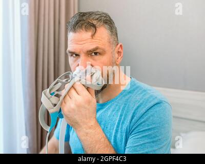 Reifer glücklicher Mann mit chronischen Atemproblemen erwägt die Verwendung CPAP-Maschine auf dem Bett im Schlafzimmer sitzen. Gesundheitswesen, CPAP, Schnarchen Konzept Stockfoto