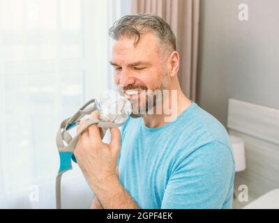 Reifer glücklicher Mann mit chronischen Atemproblemen erwägt die Verwendung CPAP-Maschine auf dem Bett im Schlafzimmer sitzen. Gesundheitswesen, CPAP, Schnarchen Konzept Stockfoto