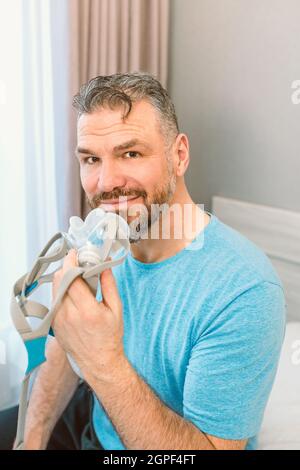 Reifer glücklicher Mann mit chronischen Atemproblemen erwägt die Verwendung CPAP-Maschine auf dem Bett im Schlafzimmer sitzen. Gesundheitswesen, CPAP, Schnarchen Konzept Stockfoto