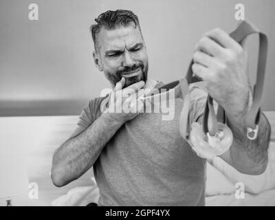 Unglücklich schockiert Mann mit chronischen Atemproblemen überrascht, indem CPAP-Maschine auf dem Bett im Schlafzimmer sitzen. Stockfoto