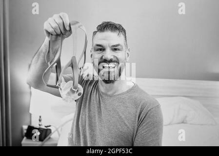 Monochromes Porträt eines glücklichen ausgeruhten Mannes mit chronischen Atemproblemen nach der Verwendung eines CPAP-Geräts, das im Schlafzimmer auf dem Bett sitzt. Gesundheitswesen, CPAP Stockfoto