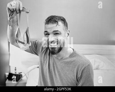 Monochromes Porträt eines glücklichen ausgeruhten Mannes mit chronischen Atemproblemen nach der Verwendung eines CPAP-Geräts, das im Schlafzimmer auf dem Bett sitzt. Gesundheitswesen, CPAP Stockfoto
