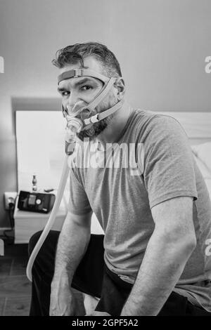 Unglücklich schockiert Mann mit chronischen Atemproblemen überrascht, indem CPAP-Maschine auf dem Bett im Schlafzimmer sitzen. Stockfoto