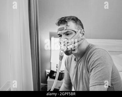 Unglücklich schockiert Mann mit chronischen Atemproblemen überrascht, indem CPAP-Maschine auf dem Bett im Schlafzimmer sitzen. Stockfoto