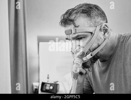 Unglücklich schockiert Mann mit chronischen Atemproblemen überrascht, indem CPAP-Maschine auf dem Bett im Schlafzimmer sitzen. Stockfoto