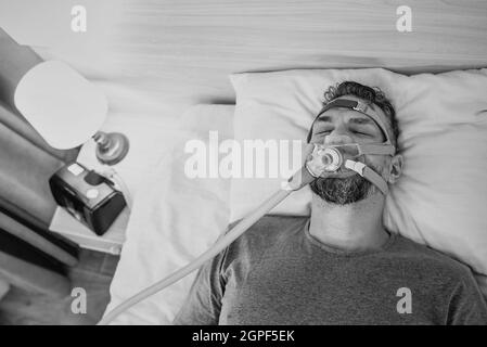 Monochromes Porträt eines schlafenden Mannes mit chronischen Atemproblemen erwägt die Verwendung des CPAP-Geräts im Bett. Gesundheitswesen, obstruktive Schlafapnoe-Therapie Stockfoto