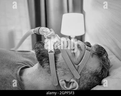 Monochromes Porträt eines schlafenden Mannes mit chronischen Atemproblemen erwägt die Verwendung des CPAP-Geräts im Bett. Gesundheitswesen, obstruktive Schlafapnoe-Therapie Stockfoto