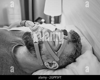 Monochromes Porträt eines schlafenden Mannes mit chronischen Atemproblemen erwägt die Verwendung des CPAP-Geräts im Bett. Gesundheitswesen, obstruktive Schlafapnoe-Therapie Stockfoto