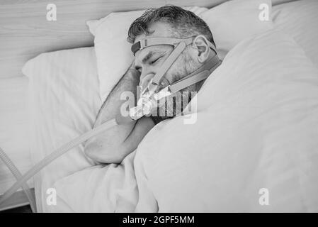 Monochromes Porträt eines schlafenden Mannes mit chronischen Atemproblemen erwägt die Verwendung des CPAP-Geräts im Bett. Gesundheitswesen, obstruktive Schlafapnoe-Therapie Stockfoto