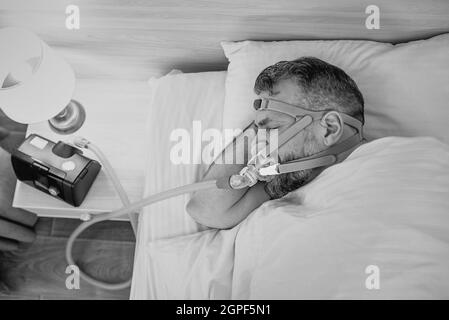 Monochromes Porträt eines schlafenden Mannes mit chronischen Atemproblemen erwägt die Verwendung des CPAP-Geräts im Bett. Gesundheitswesen, obstruktive Schlafapnoe-Therapie Stockfoto