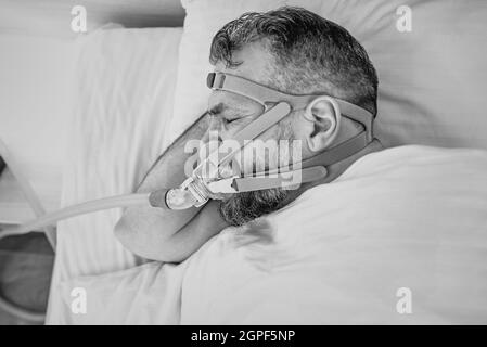 Monochromes Porträt eines schlafenden Mannes mit chronischen Atemproblemen erwägt die Verwendung des CPAP-Geräts im Bett. Gesundheitswesen, obstruktive Schlafapnoe-Therapie Stockfoto