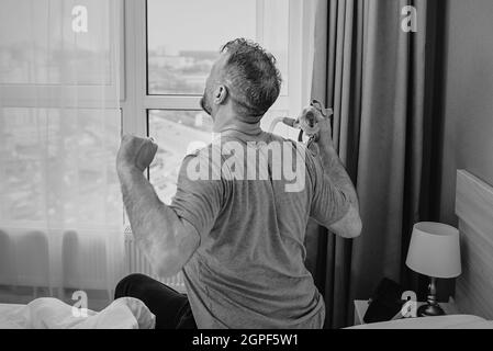 Monochromes Porträt eines glücklichen ausgeruhten Mannes mit chronischen Atemproblemen nach der Verwendung eines CPAP-Geräts, das im Schlafzimmer auf dem Bett sitzt. Gesundheitswesen, CPAP Stockfoto