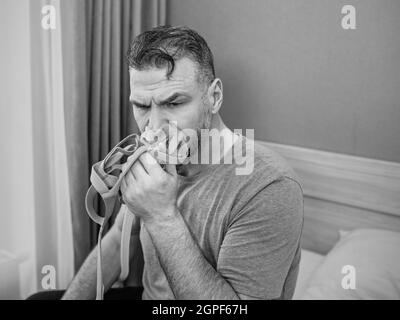 Unglücklich schockiert Mann mit chronischen Atemproblemen überrascht, indem CPAP-Maschine auf dem Bett im Schlafzimmer sitzen. Stockfoto