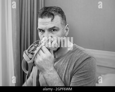 Unglücklich schockiert Mann mit chronischen Atemproblemen überrascht, indem CPAP-Maschine auf dem Bett im Schlafzimmer sitzen. Stockfoto