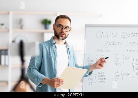Junger männlicher Lehrer, der auf eine Tafel mit englischen Grammatikregeln zeigt und von zu Hause aus Online-Unterricht durchführt Stockfoto