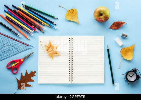Leerer Notizblock und Schulbedarf, Wecker, Apfel und gelbe Herbstblätter auf blauem Hintergrund. Zurück zum Schulkonzept. Draufsicht, flaches Lay, Mock Up Stockfoto