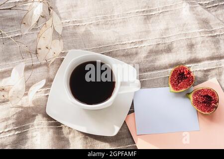 Kaffee in einer weißen Tasse, Feigen und Lunaria trockene Blume auf textilen Hintergrund. Stillleben im Herbst. Orangefarbener Umschlag mit leerer Papierkarte. Draufsicht. Stockfoto