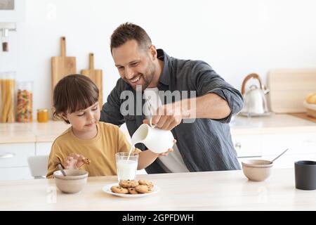 Gesunder Snack für Kinder. Glücklicher Vater gießt frische Milch für seinen Sohn, bereitet das Mittagessen für den kleinen Jungen, sitzt in der Küche Stockfoto