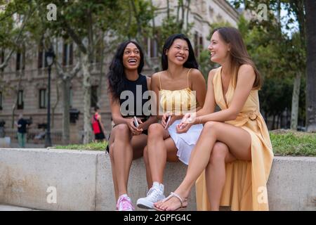 Eine Gruppe junger multirassischer Freundinnen in stilvollen Outfits, die am Rand sitzen und fröhlich lachen, während sie sich unterhalten und den Sommertag gemeinsam in der Stadt genießen Stockfoto