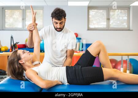 Frau, die auf dem Rücken liegt und Oberkörperübungen mit Hilfe eines männlichen Physiotherapeuten macht. Stockfoto