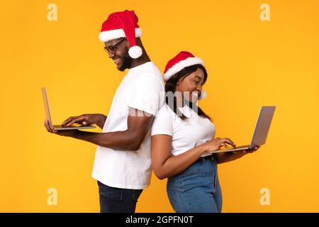 Afrikanisches Paar Mit Laptop Trägt Santa Hats Stehend, Gelber Hintergrund Stockfoto