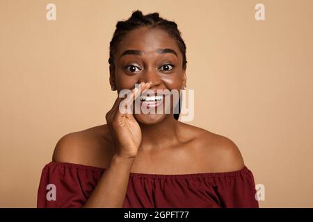 Hübsche schwarze Dame, die Gerüchte teilt und die Handfläche neben den Mund hält Stockfoto