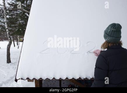 Mädchen steht mit dem Rücken und schreibt die Zahl 2022 in den Schnee. Spaziergänge am Heiligabend, Spaß, Winterferien. Flauschige Fäustlinge. Hallo Winter. Kaltes Wetter Stockfoto