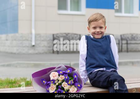 Ein süßer Erstklässler-Junge in Schuluniform mit einem schönen Blumenstrauß im Schulhof nach dem Feiertag am 1. September. Sachkenntniss Stockfoto