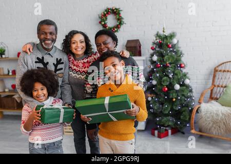 Fröhliche afroamerikanische Kinder mit weihnachtsgeschenken, die in der Nähe von Mutter und Großeltern auf die Kamera schauen Stockfoto